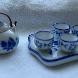 Op de afbeelding: Een miniatuur wit porseleinen theeservies met blauwe bloemmotieven. De set bevat een theepot met een bruin handvat en vier kleine kopjes op een bijpassend dienblad. De theeset heeft een delicaat, handgeschilderd ontwerp.