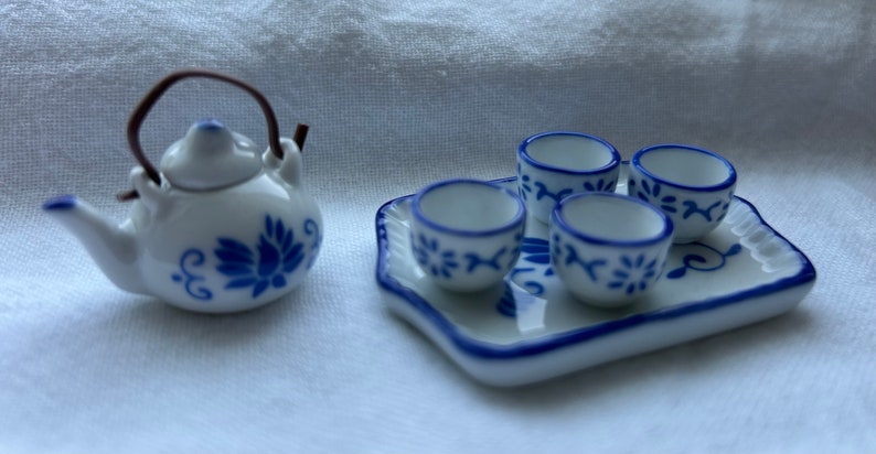 Op de afbeelding: Een miniatuur wit porseleinen theeservies met blauwe bloemmotieven. De set bevat een theepot met een bruin handvat en vier kleine kopjes op een bijpassend dienblad. De theeset heeft een delicaat, handgeschilderd ontwerp.