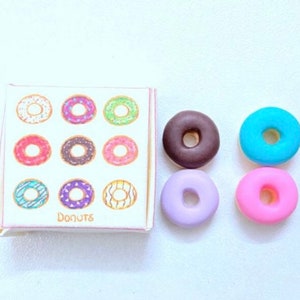 May include: A box of donuts with a variety of colorful donuts printed on the front.  The box says "Donuts".  There are four miniature donuts in different colors: brown, blue, purple, and pink.