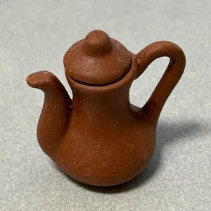 May include: A small, terracotta-colored teapot with a lid. The teapot has a rounded body and a curved spout. The handle is attached to the side of the teapot.