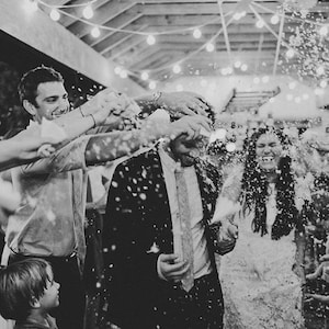 May include: A black and white photo of a wedding reception. The bride and groom are being showered with confetti by their guests. The bride is wearing a white dress and the groom is wearing a suit. The guests are smiling and laughing.