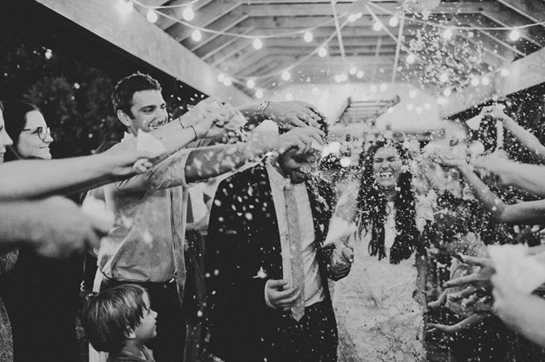 May include: A black and white photo of a wedding reception. The bride and groom are being showered with confetti by their guests. The bride is wearing a white dress and the groom is wearing a suit. The guests are smiling and laughing.