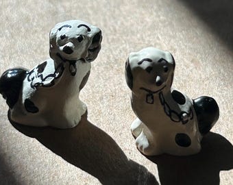 以下が含まれることがあります： 白い体に黒い斑点と絵付けが施されたセラミック製の犬の置物2体。1体は頭を傾けて座り、もう1体は頭を横に傾けて立っている。どちらも黒い首輪に鎖が付いている。
