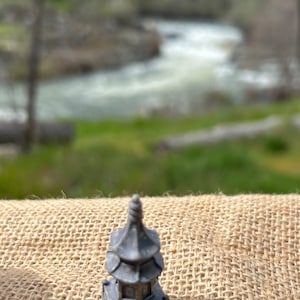 May include: A small, gray, decorative pagoda-style structure with a pointed roof and intricate details. The structure is sitting on a brown burlap surface.
