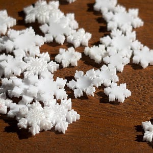 May include: A collection of small, white, plastic snowflake-shaped embellishments with intricate, six-pointed geometric patterns. Each individual piece features a smooth, glossy finish and a flat base, scattered across the frame in various orientations.