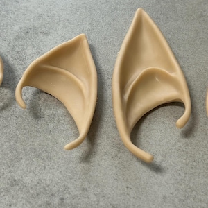 May include: Four beige elf ear prosthetics. Two pairs of pointed, curved ears are arranged on a gray surface. The ears have a smooth texture and are designed to be worn for cosplay or costume purposes.