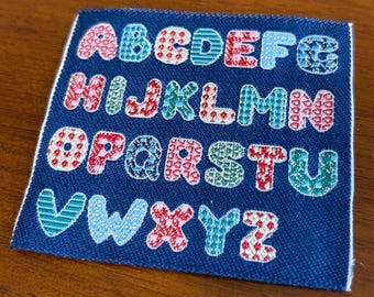 May include: A blue fabric patch with the alphabet in white outlined letters. Each letter is filled with a different colorful patterned fabric.