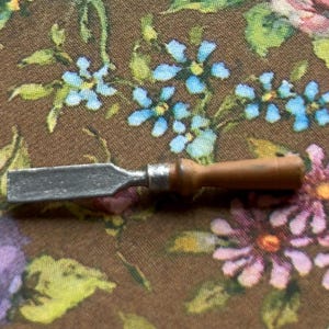 以下が含まれることがあります： 銀色の刃と茶色の木製ハンドルが付いた小さな木工用ノミ。紫、ピンク、青の花柄の背景に配置されています。木を彫ったり成形したりするために使用される可能性があります。