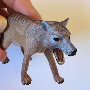 May include: A tan and brown toy thylacine, also known as a Tasmanian tiger, with black stripes on its back. The toy has an open mouth, revealing a golden interior. The thylacine is a marsupial, now extinct.