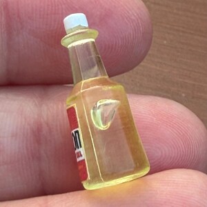Pode incluir: Uma garrafa em miniatura amarelo transl&uacute;cido com tampa branca e um r&oacute;tulo vermelho com o texto parcial 'ON'. A garrafa tem um gargalo afunilado e um corpo retangular com uma pequena protuber&acirc;ncia moldada na superf&iacute;cie frontal do objeto.