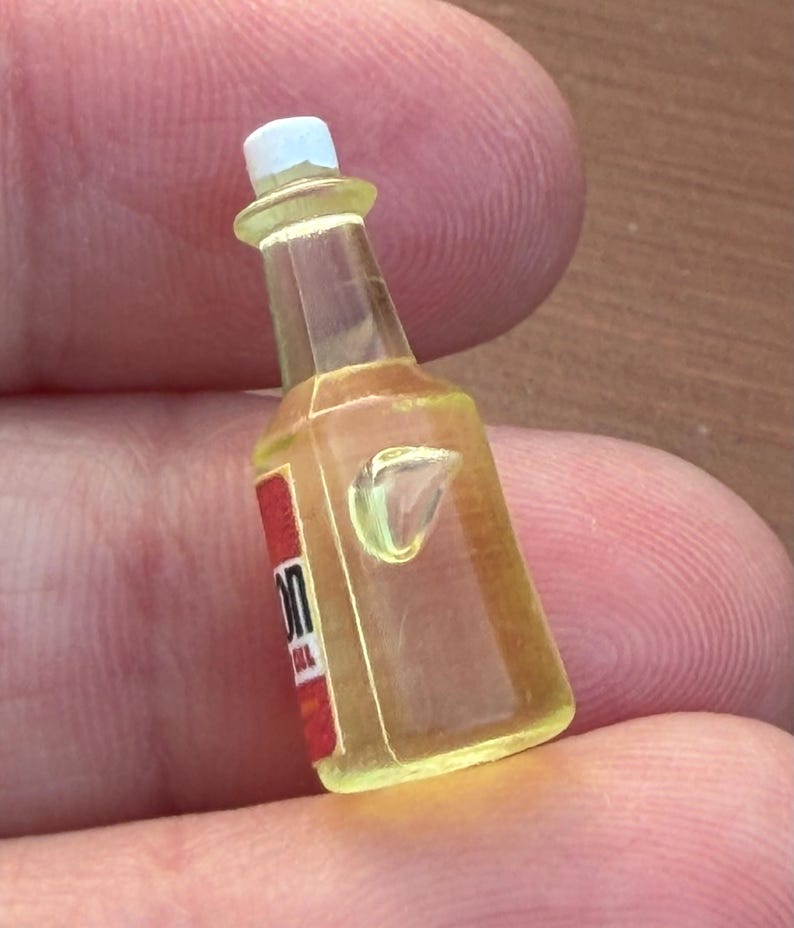 Pode incluir: Uma garrafa em miniatura amarelo transl&uacute;cido com tampa branca e um r&oacute;tulo vermelho com o texto parcial 'ON'. A garrafa tem um gargalo afunilado e um corpo retangular com uma pequena protuber&acirc;ncia moldada na superf&iacute;cie frontal do objeto.