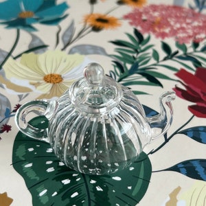 May include: A clear glass teapot with a ribbed design. The teapot has a lid and a spout. The teapot is sitting on a floral patterned surface.