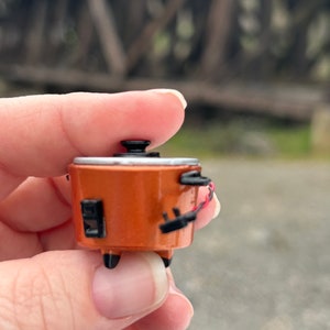 May include: A small, orange-brown slow cooker with a black lid and black accents. The slow cooker is being held in a hand.