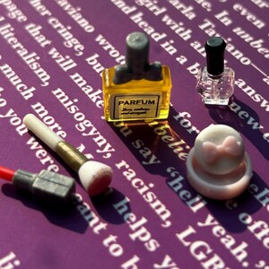 Könnte beinhalten: Ein Set an Miniatur-Schönheitsprodukten, darunter eine rosa-weiße Kompaktdose mit Schleife, ein klares Fläschchen Nagellack, ein rot-grauer Lippenstift, ein weiß-brauner Make-up-Pinsel und ein kleines Fläschchen Parfüm mit der Aufschrift "PARFUM".