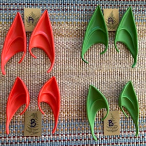 May include: A collection of elf ear prosthetics in red and green. Each pair of ears is made of a flexible material and has a curved shape. Small cards with the letters "A" and "B" are included.