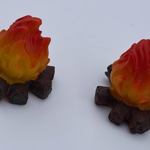 Peut inclure: Deux figurines miniatures de feu de camp composées de bûches sculptées brun foncé disposées en cercle, avec des flammes jaune vif et orange s'élevant du centre. Les flammes ont une texture lisse et brillante, tandis que les bûches sont mates.