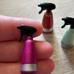 May include: A close-up of three miniature spray bottles in pink, red, and light green. Each bottle has a black spray nozzle and a silver band near the base. The pink bottle is in focus.