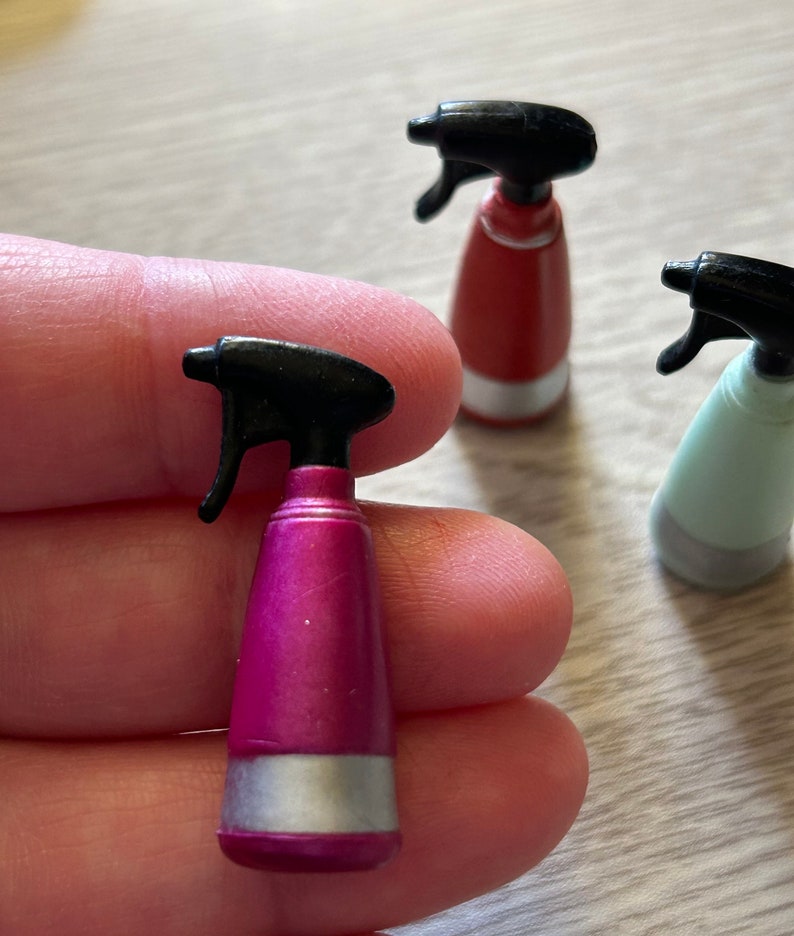 May include: A close-up of three miniature spray bottles in pink, red, and light green. Each bottle has a black spray nozzle and a silver band near the base. The pink bottle is in focus.