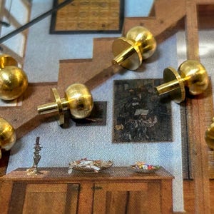 Pode incluir: Cinco botões de armário de metal dourado com topos redondos e lisos e uma base plana. Os botões estão dispostos sobre uma superfície de madeira.