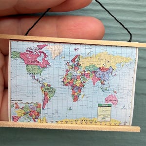 May include: A miniature wall hanging featuring a colorful world map printed on paper, secured between two thin horizontal wooden slats at the top and bottom. A thin black string is attached to the top wooden slat for hanging purposes.