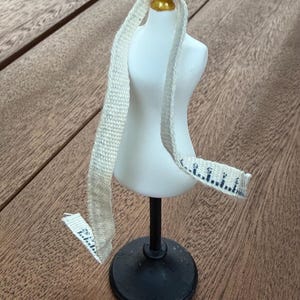 May include: A white miniature dress form with a gold-colored neck topper, mounted on a black stand. A beige fabric measuring tape with black markings is draped over the shoulders of the form, showing measurements in inches up to 52 inches.