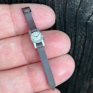 May include: A miniature silver wristwatch with a white face and black leather strap.