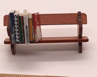 May include: A miniature wooden bench with a brown finish, featuring a set of miniature books stacked on the seat. The books have colorful covers and one has the word "ROBINSON" printed on the spine.