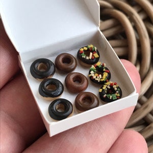 May include: A miniature box of donuts. The donuts are in a white box and have a variety of toppings. Some are glazed with chocolate, others with sprinkles. The donuts are small, and the box is held in a hand.