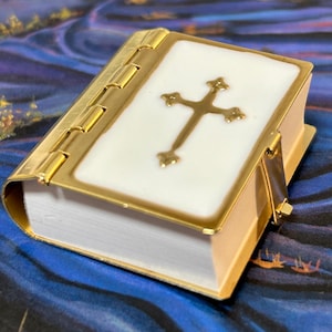 May include: A small, white porcelain book with a gold cross on the cover. The book has a gold hinge and a gold clasp.