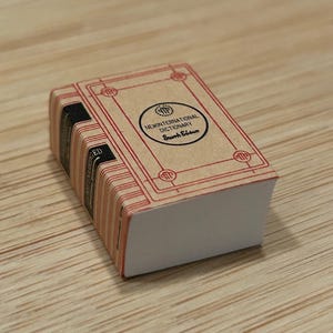 Puede incluir: Un libro en miniatura de color marrón y rojo con el título "New International Dictionary, Seventh Edition" en la portada.
