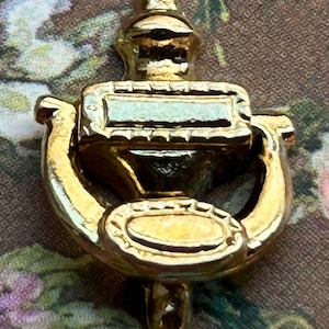 Könnte beinhalten: Ein goldfarbener Türklopfer mit klassischem Design. Der Klopfer hat eine rechteckige Platte, einen gebogenen Griff und eine ovale Schlagplatte. Das Metall hat eine glänzende Oberfläche, die das Licht reflektiert.