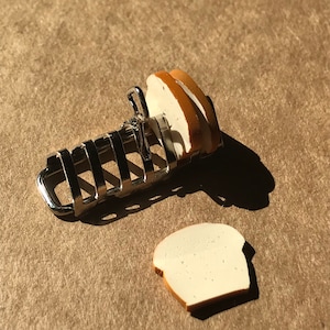 Puede incluir: Un estante para pan de metal plateado con dos rebanadas de pan blanco en la parte superior. El estante para pan está sobre una superficie marrón.