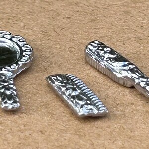 May include: A miniature silver-toned set consisting of a handheld mirror with an ornate circular frame, a small rectangular comb, and a hairbrush with a textured handle. Each piece features intricate embossed detailing on the metallic surface.