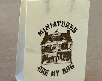 May include: A white paper bag with a brown rope handle. The bag has a brown graphic of a house with the text "Miniatures are my bag" printed on it.