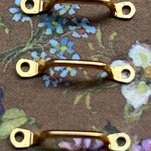 Może przedstawiać: Trzy uchwyty z metalu w kolorze złotym, każdy z zakrzywionym prętem i dwoma otworami montażowymi. Uchwyty są pokazane na tle kwiatowym z niebieskimi, fioletowymi i białymi kwiatami na brązowej tkaninie.