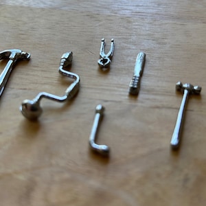 May include: A set of seven miniature silver tools, including a hammer, a wrench, a screwdriver, a pliers, and a drill. The tools are arranged on a wooden surface.