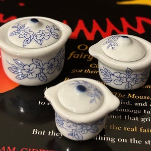 Può includere: Tre teiere in ceramica bianca con motivi floreali blu. Le teiere sono di dimensioni diverse e sono disposte su un libro con testo sulle fate.