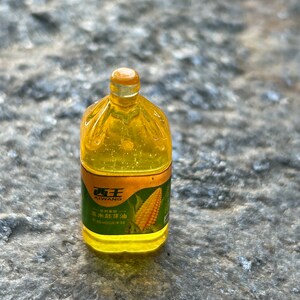 May include: A clear plastic bottle of Xi Wang brand corn oil with a yellow cap. The label is gold with green text and a picture of corn. The text on the label reads "Xi Wang" and "Corn Oil".