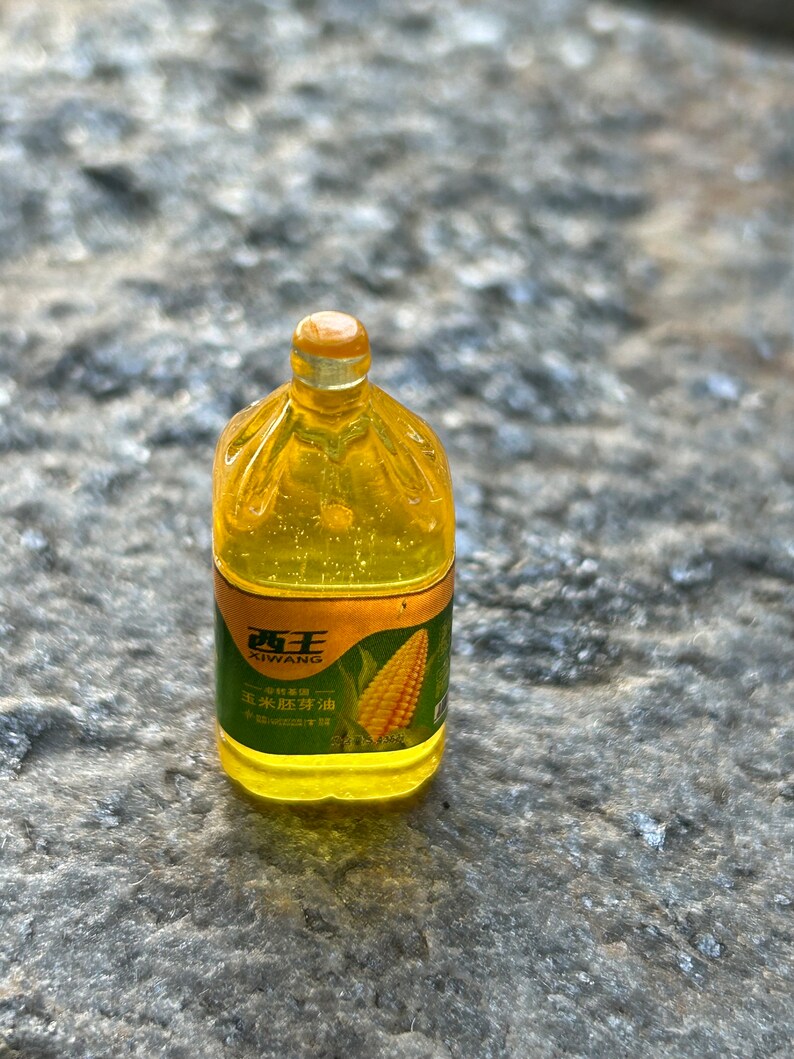 May include: A clear plastic bottle of Xi Wang brand corn oil with a yellow cap. The label is gold with green text and a picture of corn. The text on the label reads "Xi Wang" and "Corn Oil".