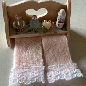 May include: A wooden shelf with a heart-shaped cutout, decorated with two glass bottles, a heart-shaped trinket, and a bottle of baby lotion. Two pink towels with white lace trim hang below the shelf.