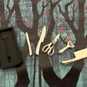 Puede incluir: Un juego de cuchillos de cocina en miniatura, un par de tijeras y un pelador, todos en plata, dispuestos sobre un fondo negro con una superficie estampada en azul oscuro y rojo.