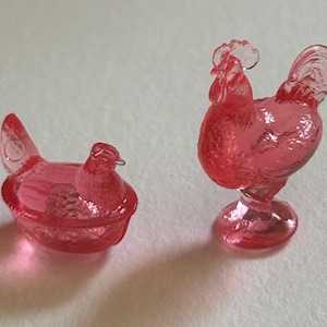 May include: Two pink glass figurines: a hen sitting in a nest and a rooster standing upright. The hen is in a sitting position, while the rooster has a detailed comb and tail. Both are made of translucent pink glass.