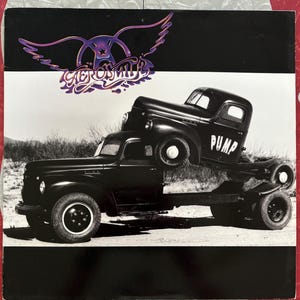 May include: A black and white photo of a vintage truck with the word "Pump" on the side. The truck is being lifted by another truck. The band Aerosmith's logo is in the top left corner of the image.