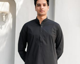 Op de afbeelding: Een man draagt een zwart lang mouw shirt met een knoopsluiting en een zwarte broek. Hij staat voor een witte muur met twee witte lampen.