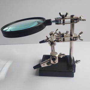 May include: A black and silver magnifying glass with a stand. The magnifying glass has a large, round lens and is attached to a flexible arm. The stand has adjustable clips and a sturdy base.