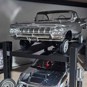 May include: A silver model car sits atop a black car lift, with another silver car below. The top car has a license plate that reads "FLOYD". A spare tire is visible on the left.