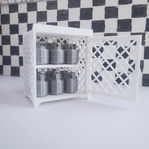 May include: A white, latticed cabinet with two shelves, each holding several silver-colored canisters. The cabinet door is open, revealing the contents. The background features a black and white checkered pattern and a brick wall.