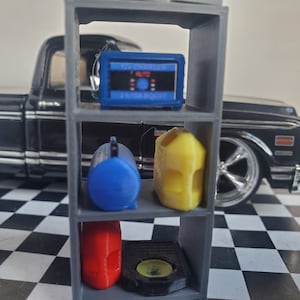 May include: A gray metal shelf unit with three shelves. The top shelf has a blue and black battery charger with the text "RV CHARGER" and "AUTO" on it. The middle shelf has a blue air compressor and a yellow gas can. The bottom shelf has a red gas can and a black round object.