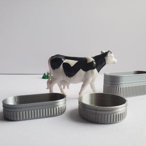 May include: A black and white plastic toy cow figurine standing on a white surface. There are three silver metal troughs in the image.