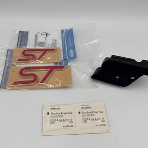 May include: Two red and silver ST emblems in clear plastic packaging, a black mounting bracket with two silver screws, and two white McKesson Alcohol Prep Pad packets measuring 1.2 inches by 2.6 inches each for surface preparation.